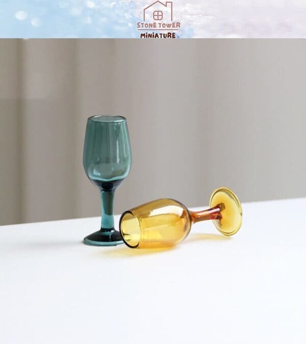 Two miniature wine glasses, one upright in teal and one lying down in amber, on a white surface.