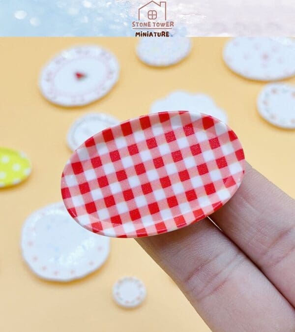 Finger holding a miniature red-checkered oval plate, with various small plates in the background on a yellow surface.