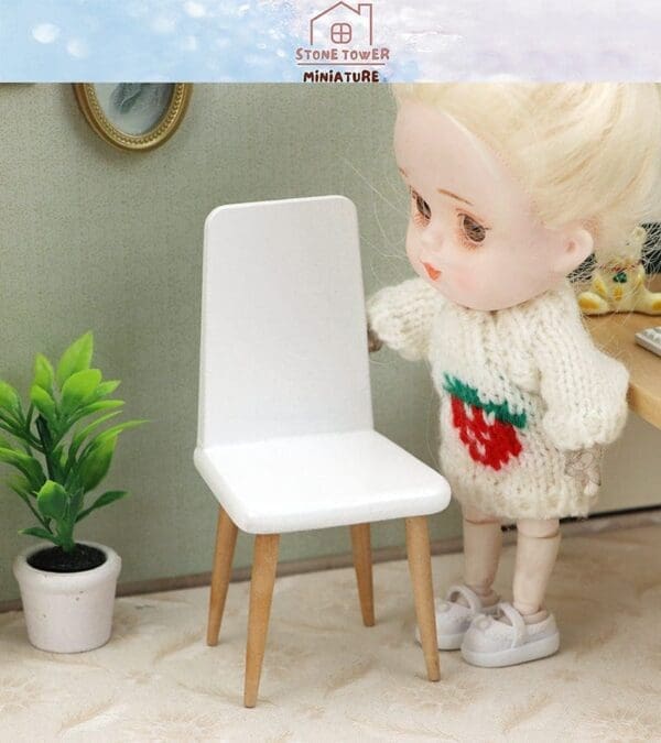 Doll in a knitted sweater stands next to a miniature white chair, with a small potted plant nearby.