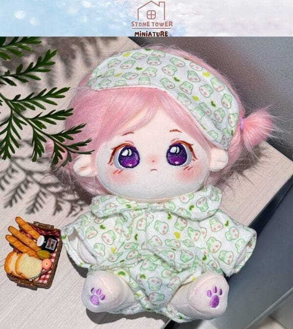 Cute plush doll with pink hair and green pajamas, sitting next to a miniature picnic basket on a light wooden surface.