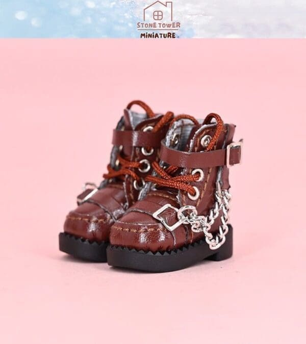 Miniature brown boots with red laces, metal eyelets, and chains, set on a pink background.