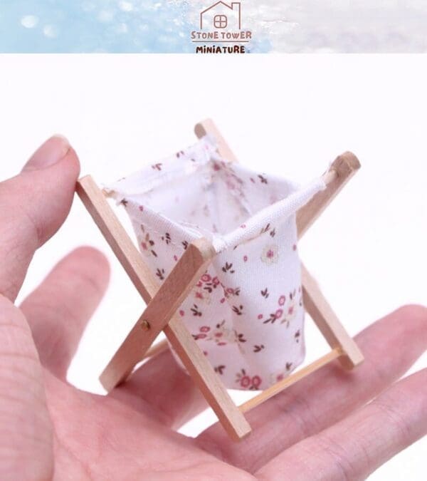 A hand holding a miniature fabric laundry basket with a floral pattern on a wooden frame.