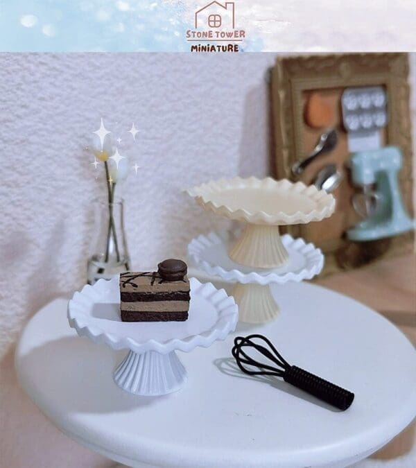 Miniature dessert on a white cake stand with a whisk, next to an empty stand and framed kitchen tools.