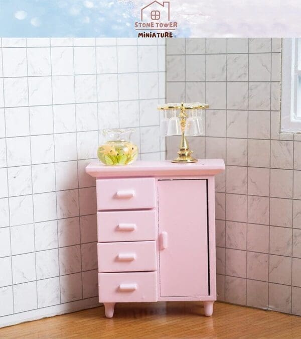 Miniature pink cabinet with drawers and a door, topped with a plant vase and a gold drink holder, in a tiled corner.