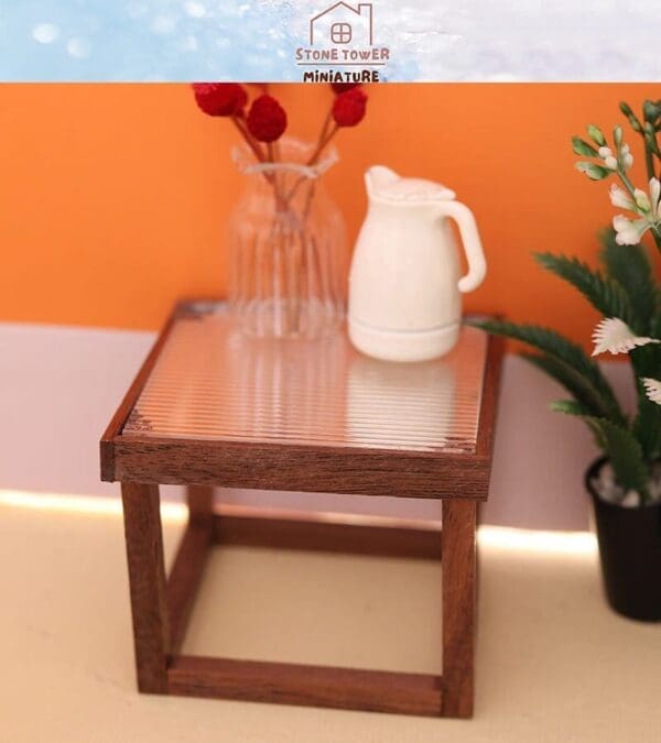 Miniature wooden table with a vase of red flowers and a white pitcher, set against an orange wall with a potted plant.