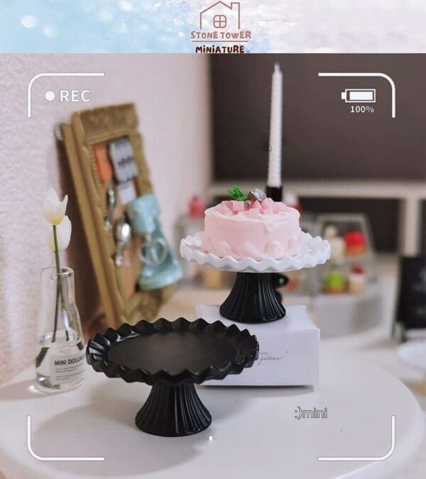 Miniature pink cake on a white stand, beside a black stand, with a small vase on a table in a cozy setting.