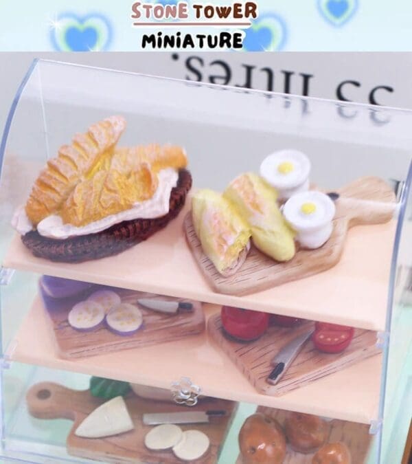 Miniature food display with croissants, eggs, and cheese on cutting boards in a clear case, labeled "Stone Tower Miniature.