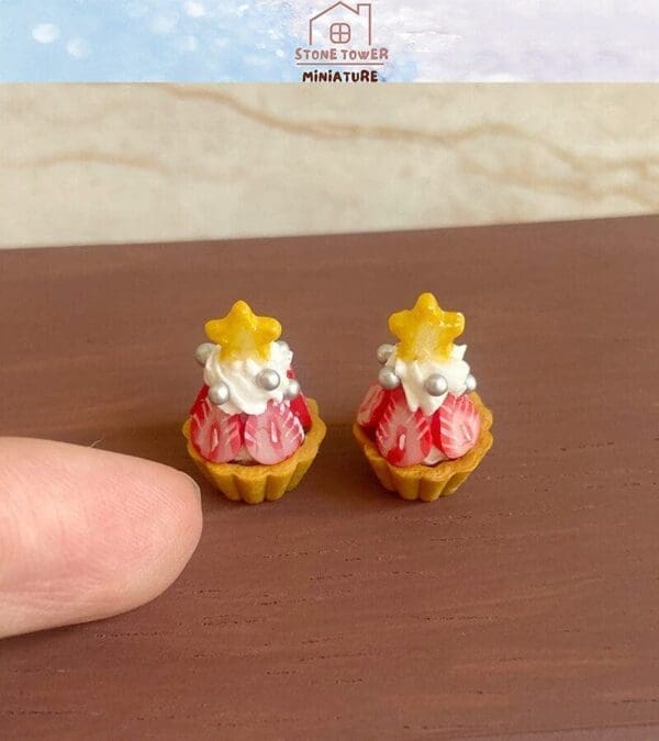 Miniature festive cupcakes with whipped cream, star toppers, and strawberry details beside a fingertip for scale.