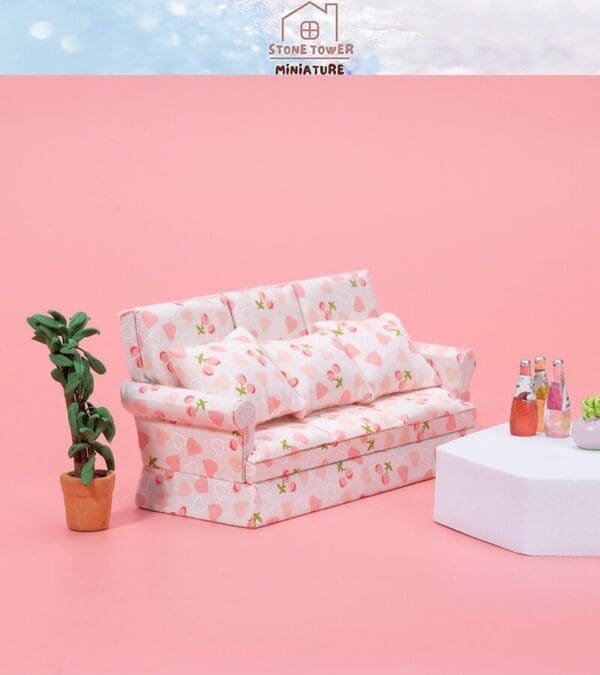 Miniature pink sofa with strawberry pattern, plant in pot, and white table with drinks on a pink background.