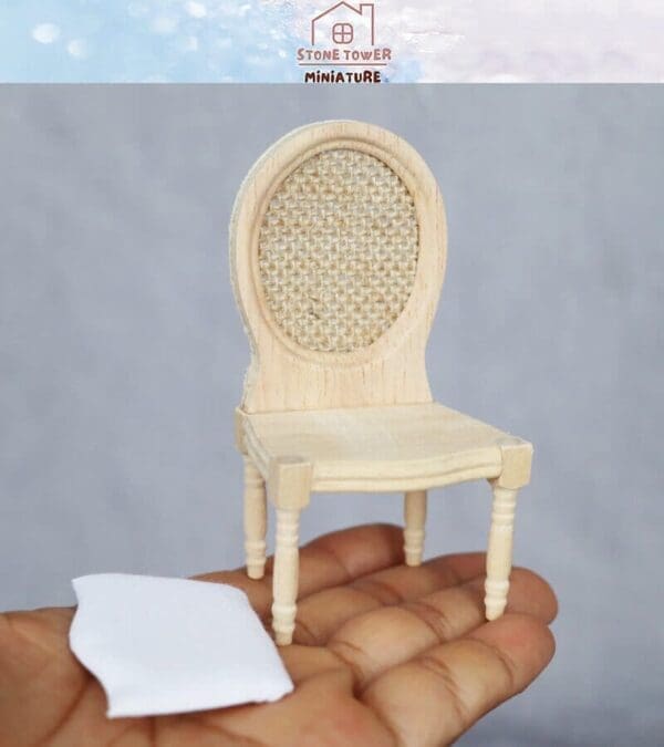 Miniature wooden chair with a woven back, held in a hand, accompanied by a small white fabric piece.