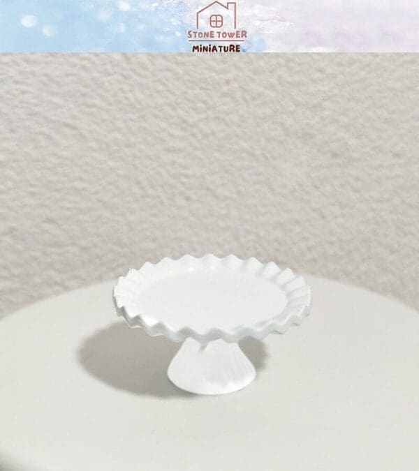 White miniature cake stand with ruffled edge on a neutral surface, logo in top corner.