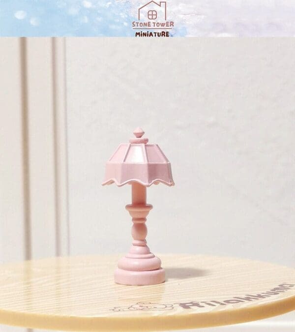 Pink miniature table lamp with a scalloped shade on a light-colored surface. Logo and text above.