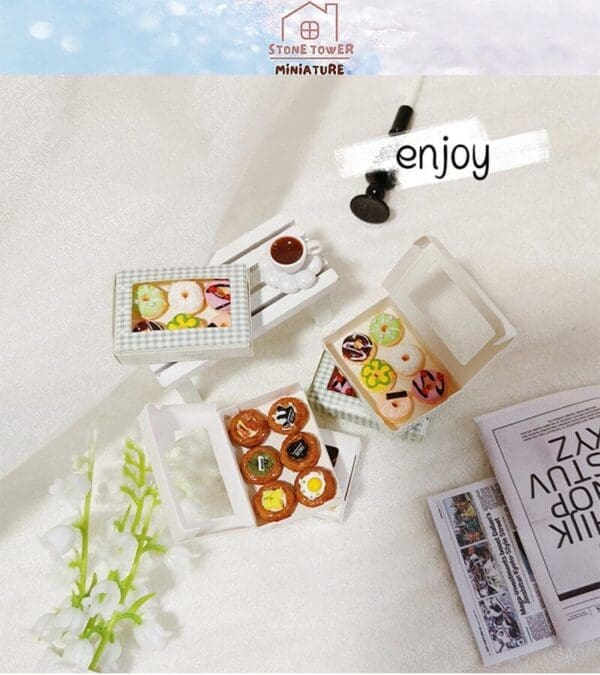 Miniature donuts in open boxes next to a tiny coffee cup and newspapers on a white surface with flowers.