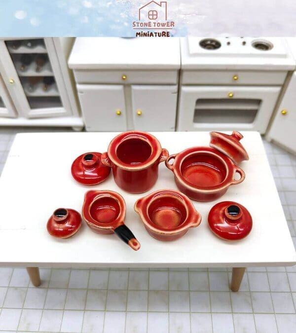 Miniature red ceramic cookware set on a white table in a model kitchen setting.