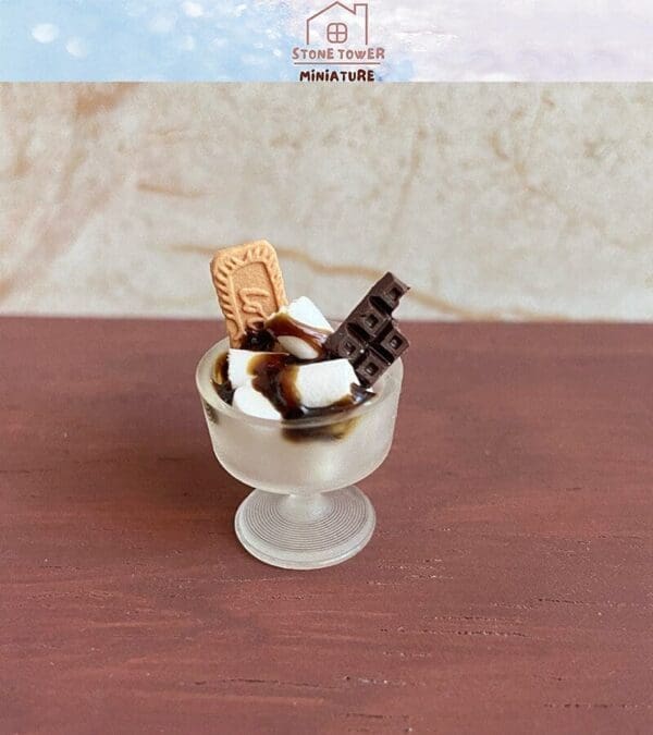 Miniature dessert in a glass features marshmallows, a biscuit, and chocolate, topped with syrup on a brown surface.