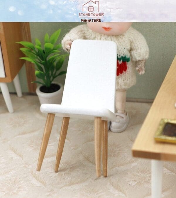 Miniature white chair with wooden legs on a floral-patterned surface, next to a doll and potted plant.