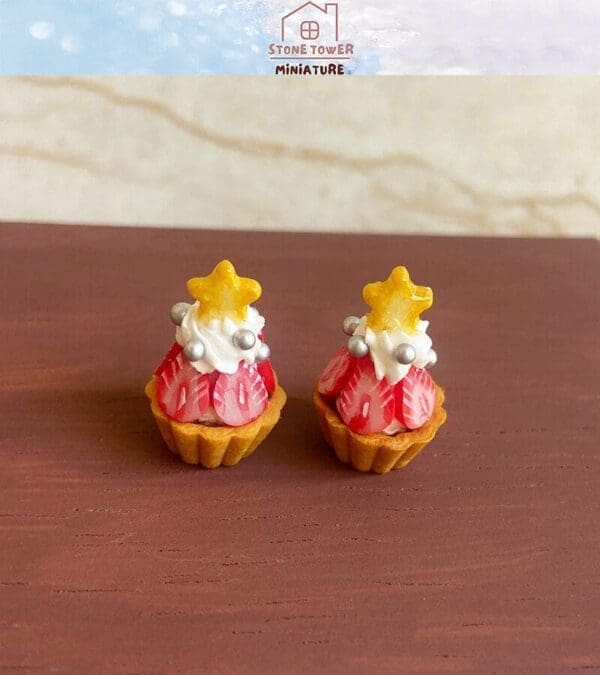 Two miniature strawberry tarts with cream, silver balls, and yellow star toppers displayed on a wooden surface.