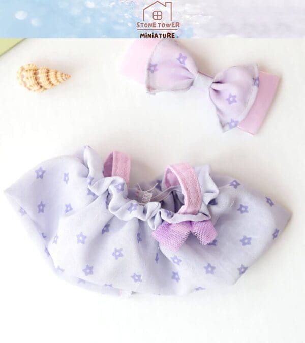 Miniature dress and bow with purple floral pattern on a white background, next to two small seashells.