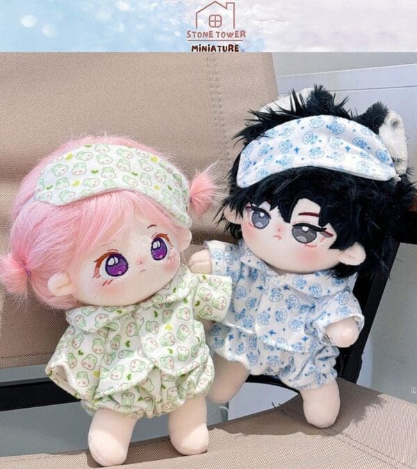 Two cute plush dolls in pajamas and sleeping masks, one with pink hair and one with black, sitting on a beige chair.