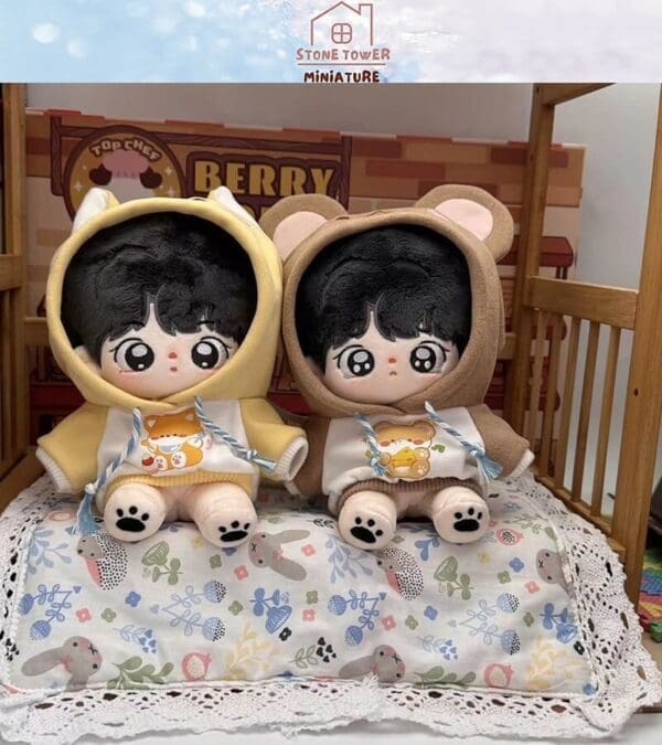 Two plush dolls in bear hoodies sit on a colorful, patterned pillow in a wooden crib setting.