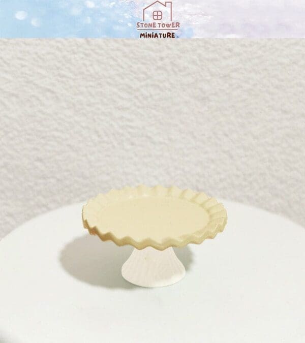 Miniature cream-colored scalloped cake stand on a round white surface, with decorative brand text above.