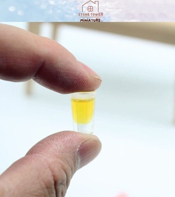 Fingers holding a tiny clear cup filled with yellow liquid, labeled "Stone Tower Miniature.