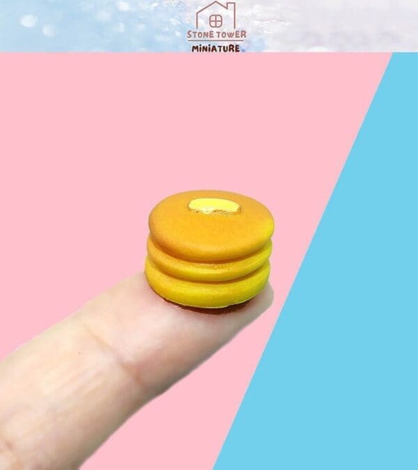 Tiny pancake stack with butter detail on a fingertip, against a pink and blue background.