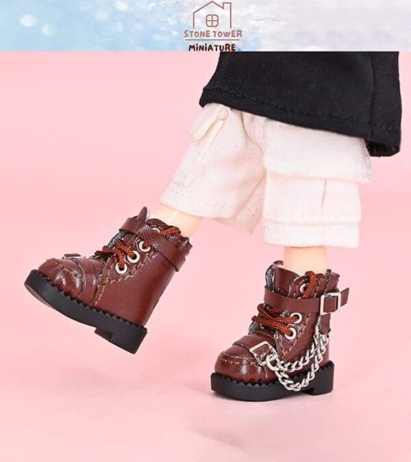 Miniature brown leather boots with red laces and chains on a doll wearing white pants, against a pink background.