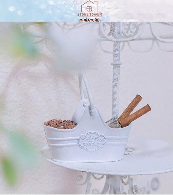 White miniature gardening bucket with soil and tools, set on an ornate metal stand against a light background.