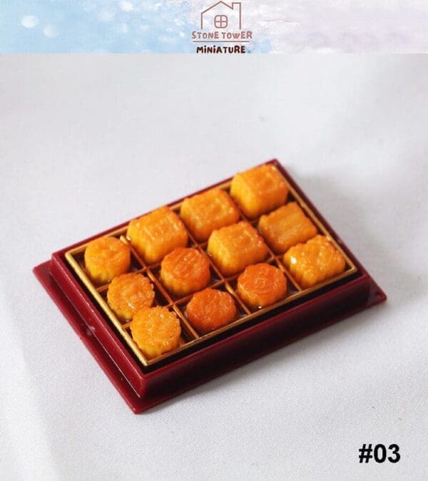 Miniature tray with 12 small, orange pastries arranged in rows on a red base, set on a white surface. #03 displayed.