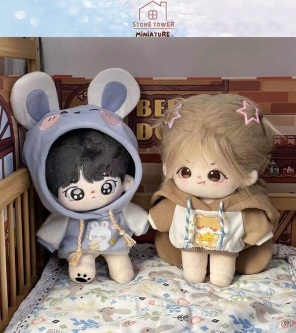 Two cute plush toys sit in a wooden crib, wearing colorful hoodies with animal designs, against a cozy backdrop.