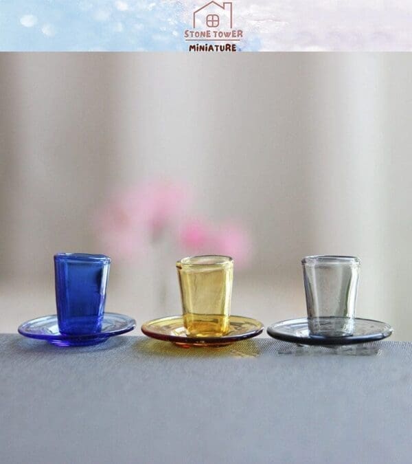 Three miniature glass cups on saucers in blue, yellow, and clear shades, displayed on a soft surface.