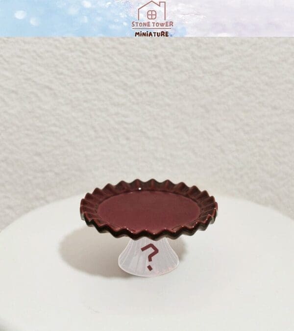 Miniature brown ruffled plate on a white stand with a question mark, placed on a light surface.