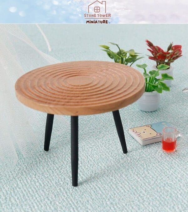 Miniature round wooden table with black legs on a textured surface, surrounded by small plants, a book, and a cup.