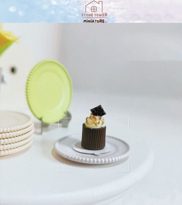 Miniature chocolate cupcake with cream and chocolate piece on a small plate, with stack of plates in the background.