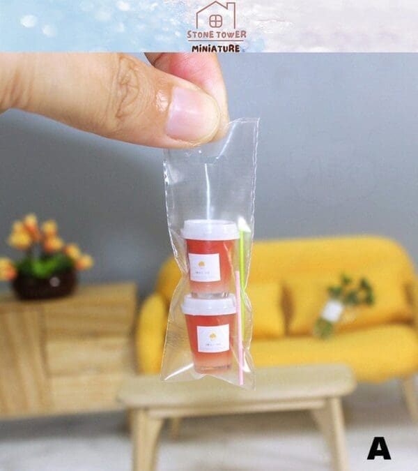 Hand holding mini orange beverage cups in a clear bag, with a yellow couch and table in the background.