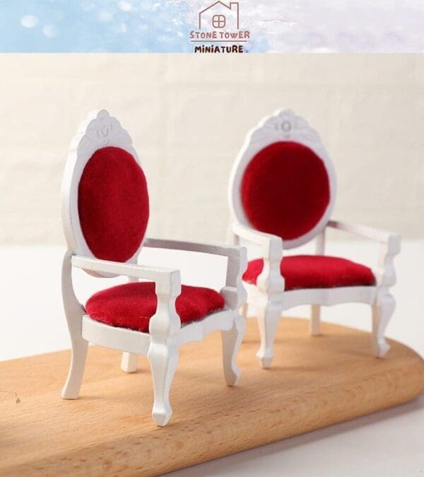 Two miniature white chairs with red velvet seats and backrests on a wooden surface.