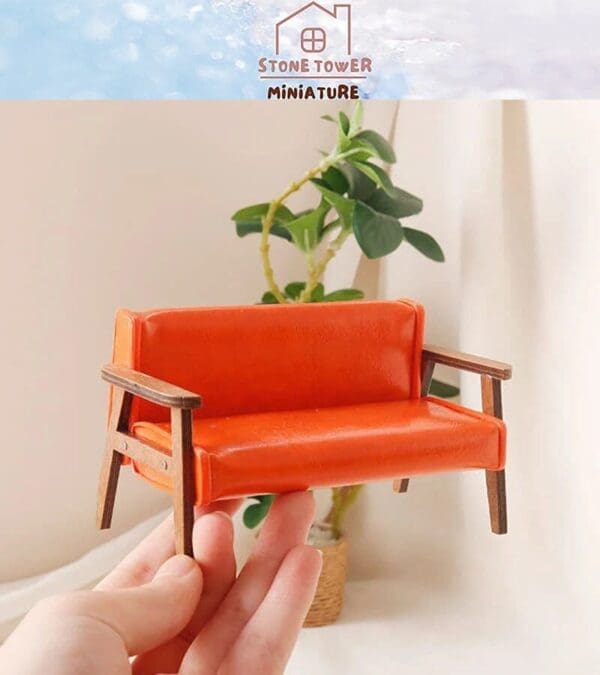 Hand holding a miniature orange sofa with wooden arms, in front of a plant and white curtains.