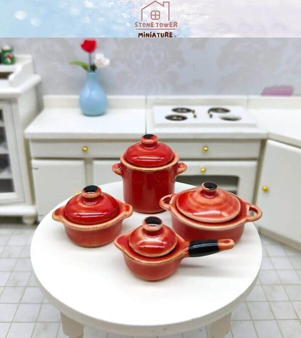 Miniature red cookware set on a white round table in a dollhouse kitchen with a blue vase and flowers.