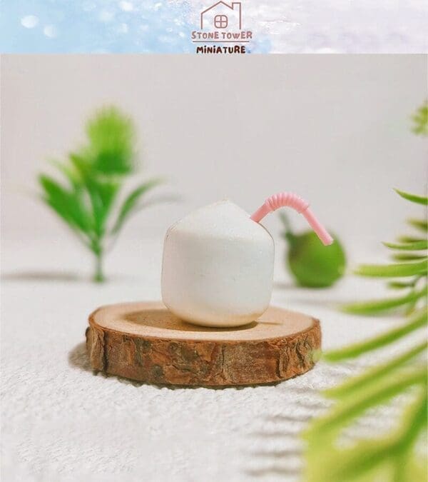 Miniature coconut with pink straw on a wooden base, surrounded by green plants with a "Stone Tower Miniature" logo above.