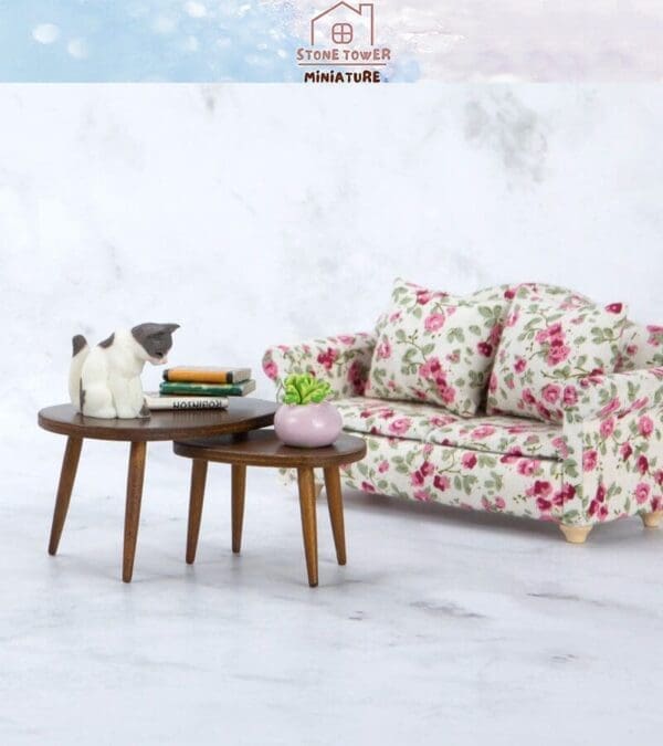 Miniature floral sofa with a cat figurine on wooden tables, books, and a small plant on a marble-look background.