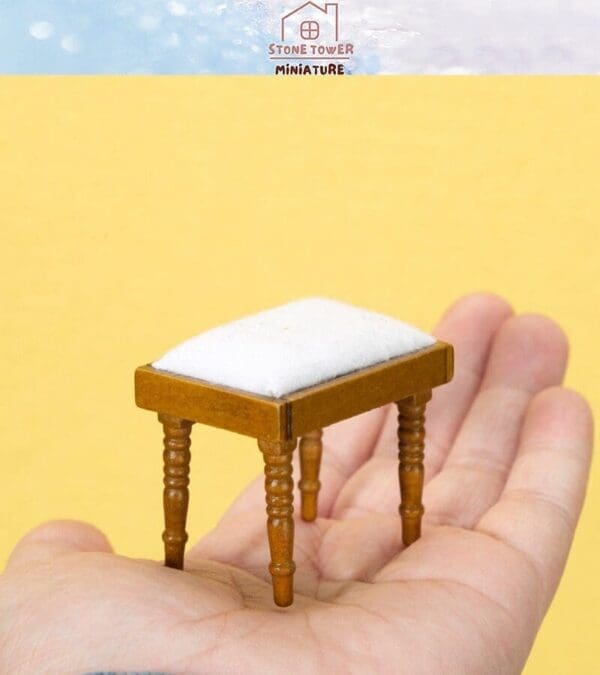 Hand holding a miniature wooden stool with a white cushion against a yellow background.