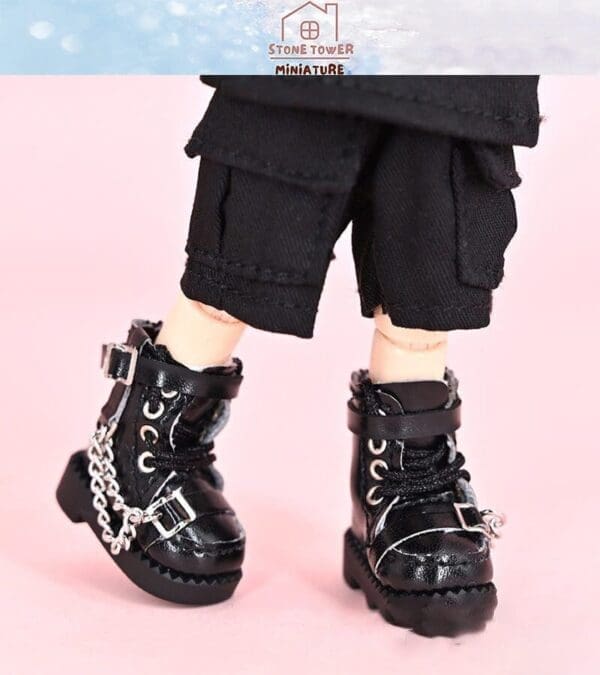 Close-up of a doll wearing black combat boots with chains and black cargo pants, on a pink background.