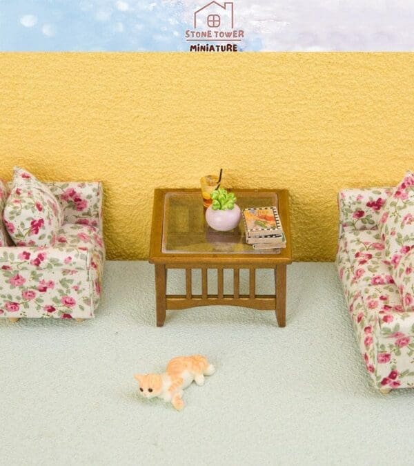 Miniature living room set with floral sofas, wooden table, small cat figure, plant, and books on a pale carpet background.