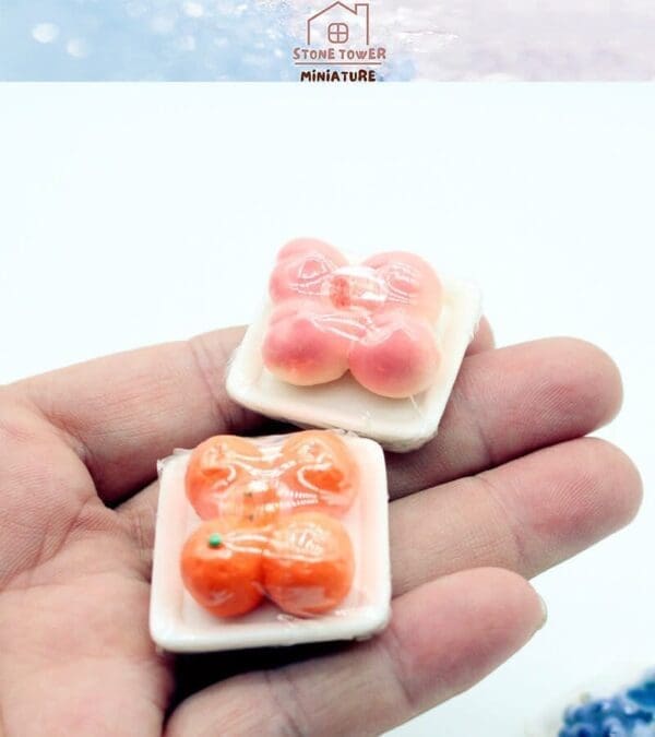 Two miniature fruit soaps, resembling peaches and oranges, on small plates held in a person's hand.