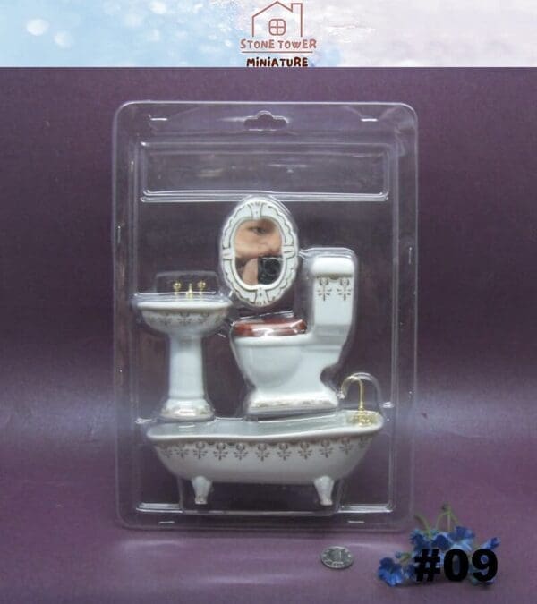 Miniature bathroom set with a sink, toilet, and tub in packaging. A face is seen in the mirror reflection.