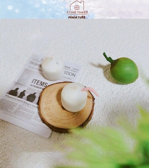 Miniature white coconuts and green mango with straw on wood slice, newspaper, and white textured surface.