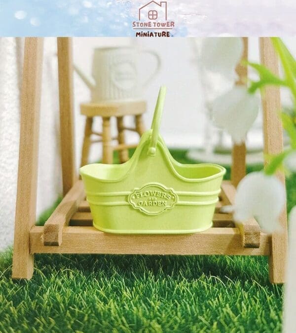 Miniature green gardening basket on wooden frame with grass, labeled "Flowers Garden," near a watering can.