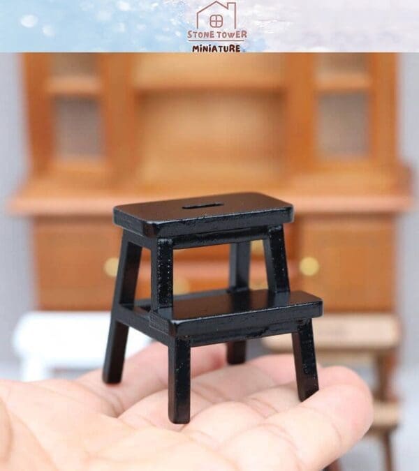 Miniature black step stool held in a hand with a wooden cabinet in the background.