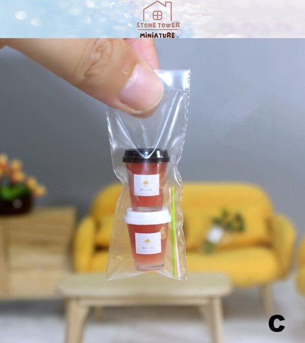 Miniature coffee cups in a clear plastic bag held by fingers, with a living room background and labeled "Stone Tower Miniature.